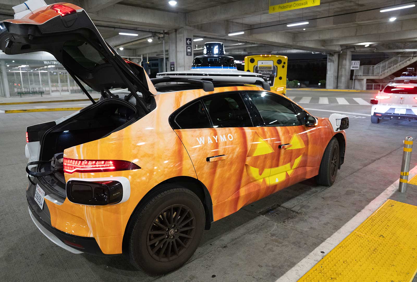 Orange Waymo autonomous vehicle in a parking deck with open trunk.